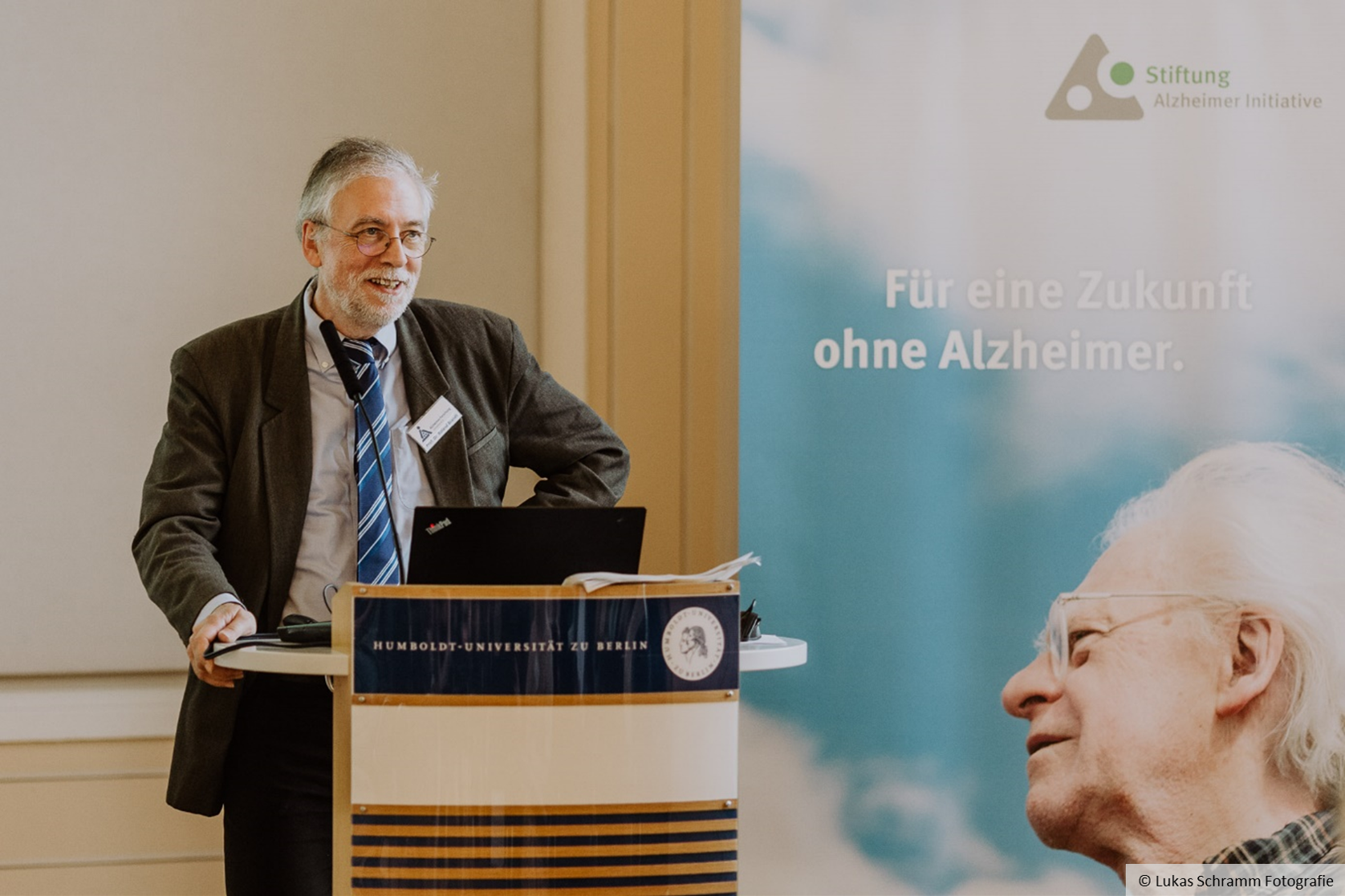 Roland Brandt stands at a lectern, next to him is a poster with the words ‘Für eine Zukunft ohne Alzheimer’.
