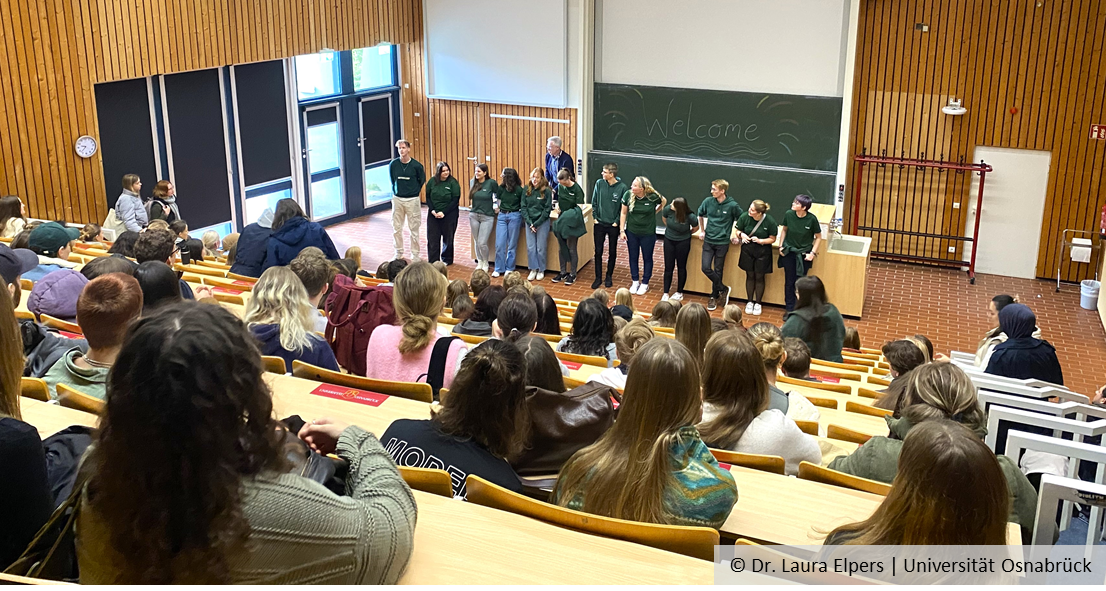 View from behind into a lecture theatre full of students. At the front are members of the student council and the Dean of Studies, Prof Michael Hensel. ‘Welcome’ is written on a blackboard.