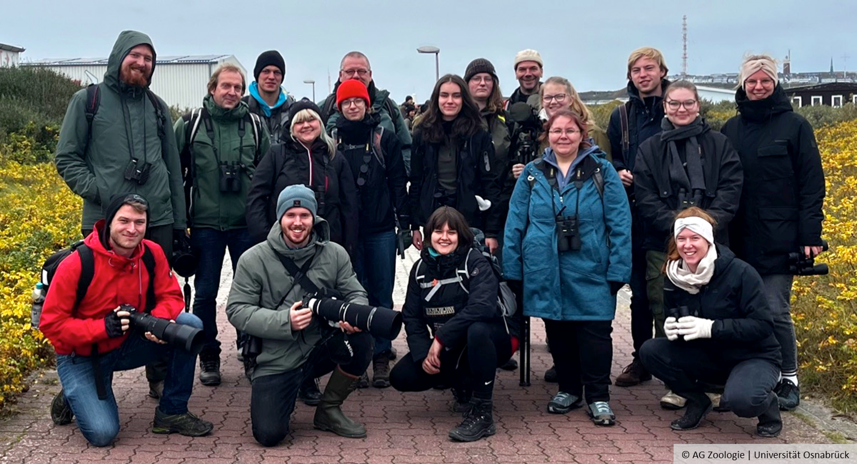 A group of people stand outside wearing jackets and smiling. Some are carrying cameras and binoculars.