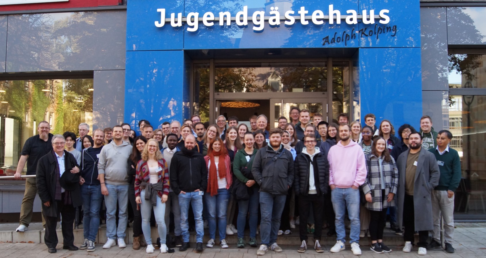 A large group of people is standing in front of a building. The words ‘Jugendgästehaus Adolph Kolping’ can be read above the entrance to the building.