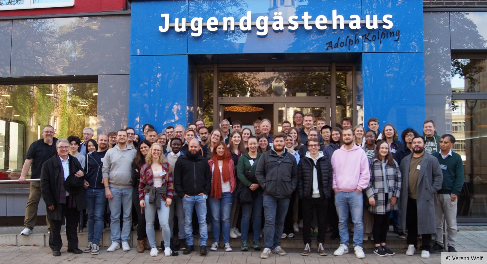 A large group of people is standing in front of a building. The words ‘Jugendgästehaus Adolph Kolping’ can be read above the entrance to the building.