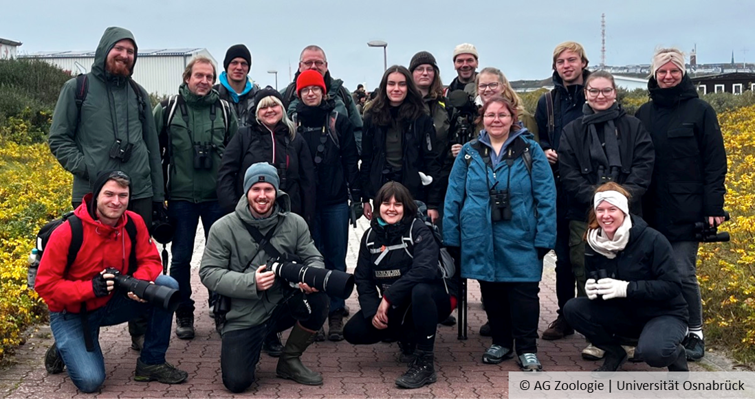 A group of people stand outside wearing jackets and smiling. Some are carrying cameras and binoculars.