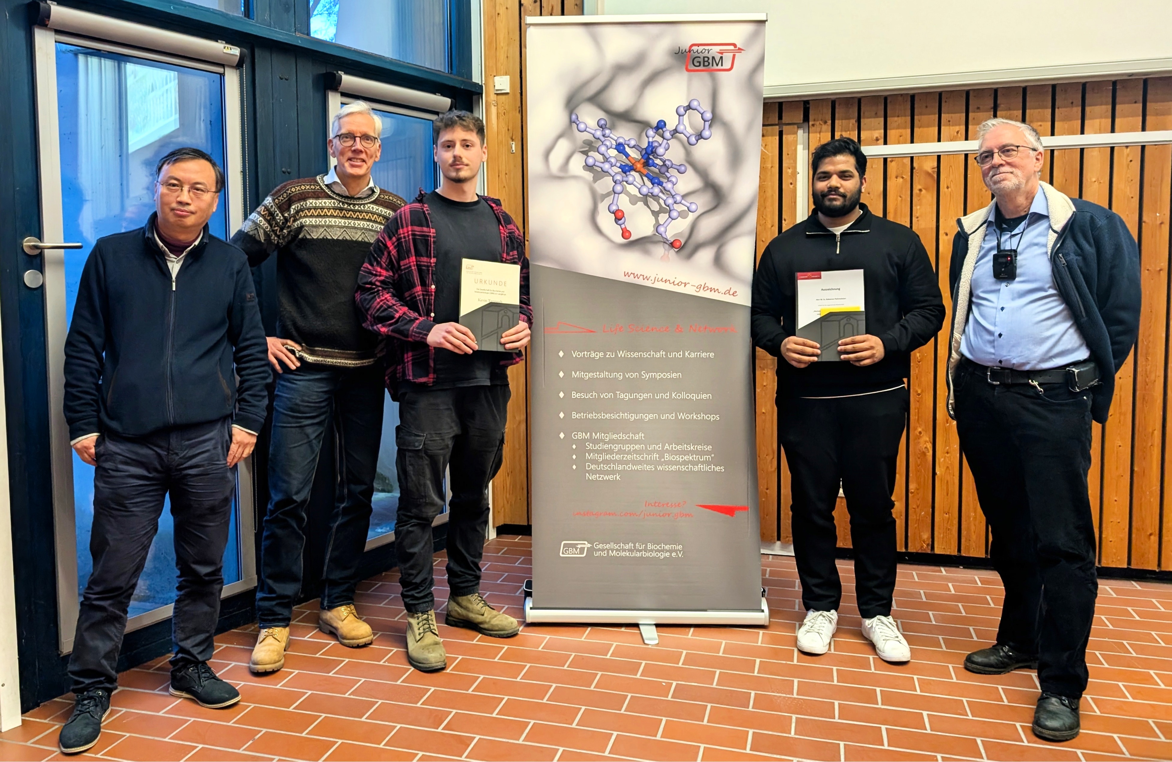 Five people smile into the camera. The two in the centre hold certificates in their hands.