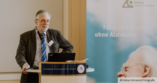 Roland Brandt is standing at a lectern, next to him is a poster with the words ‘Für eine Zukunft ohne Alzheimer’.