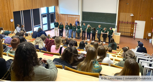 Blick von hinten in einen Hörsaal voller Studierender. Vorne stehen Mitglieder der Fachschaft und der Studiendekan Prof. Michael Hensel. An einer Tafel steht "Welcome".